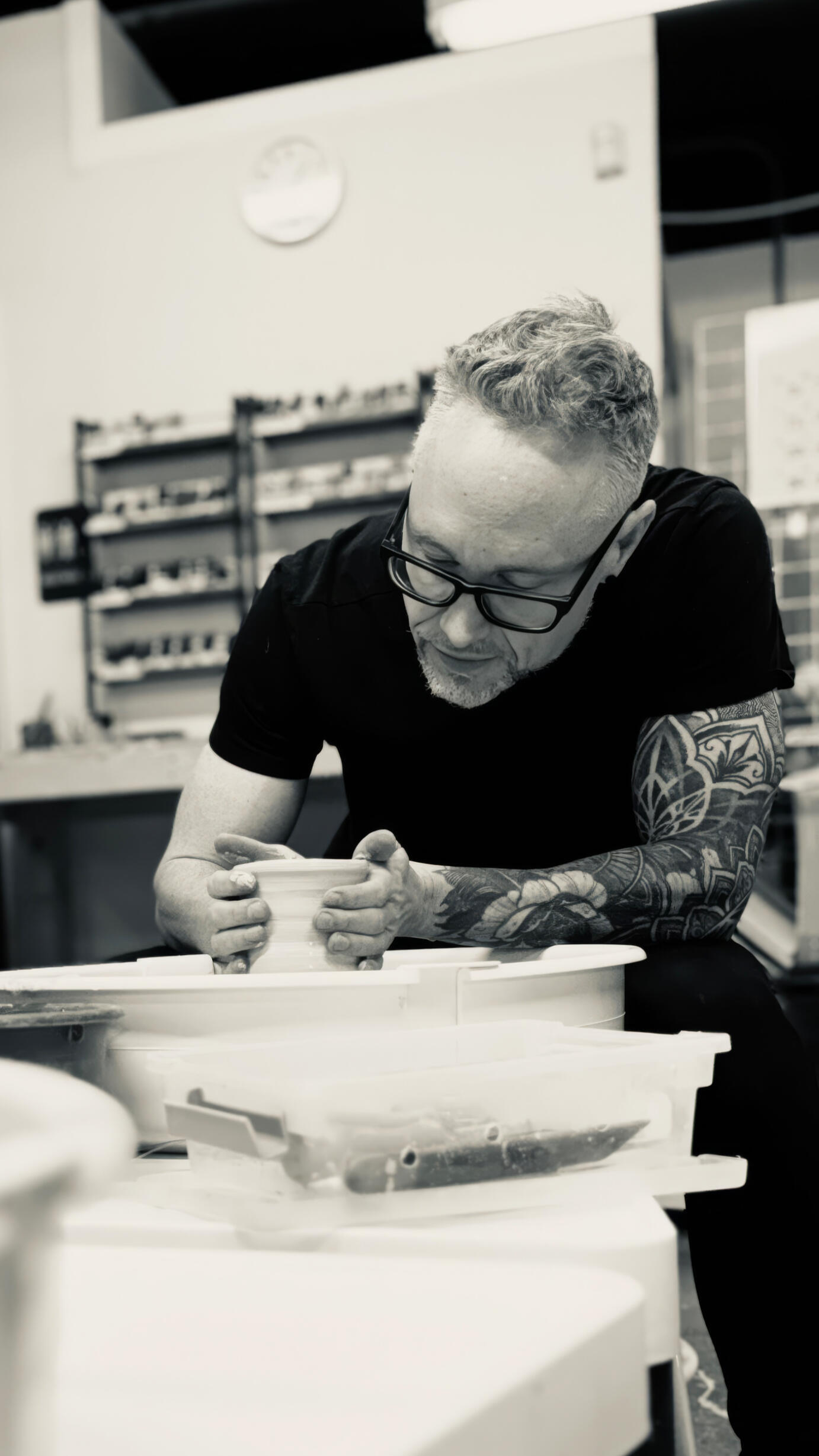 Josh Woll sculpting pottery Man with tattoo sleeve and black rim glasses shaping clay with wheel and pottery